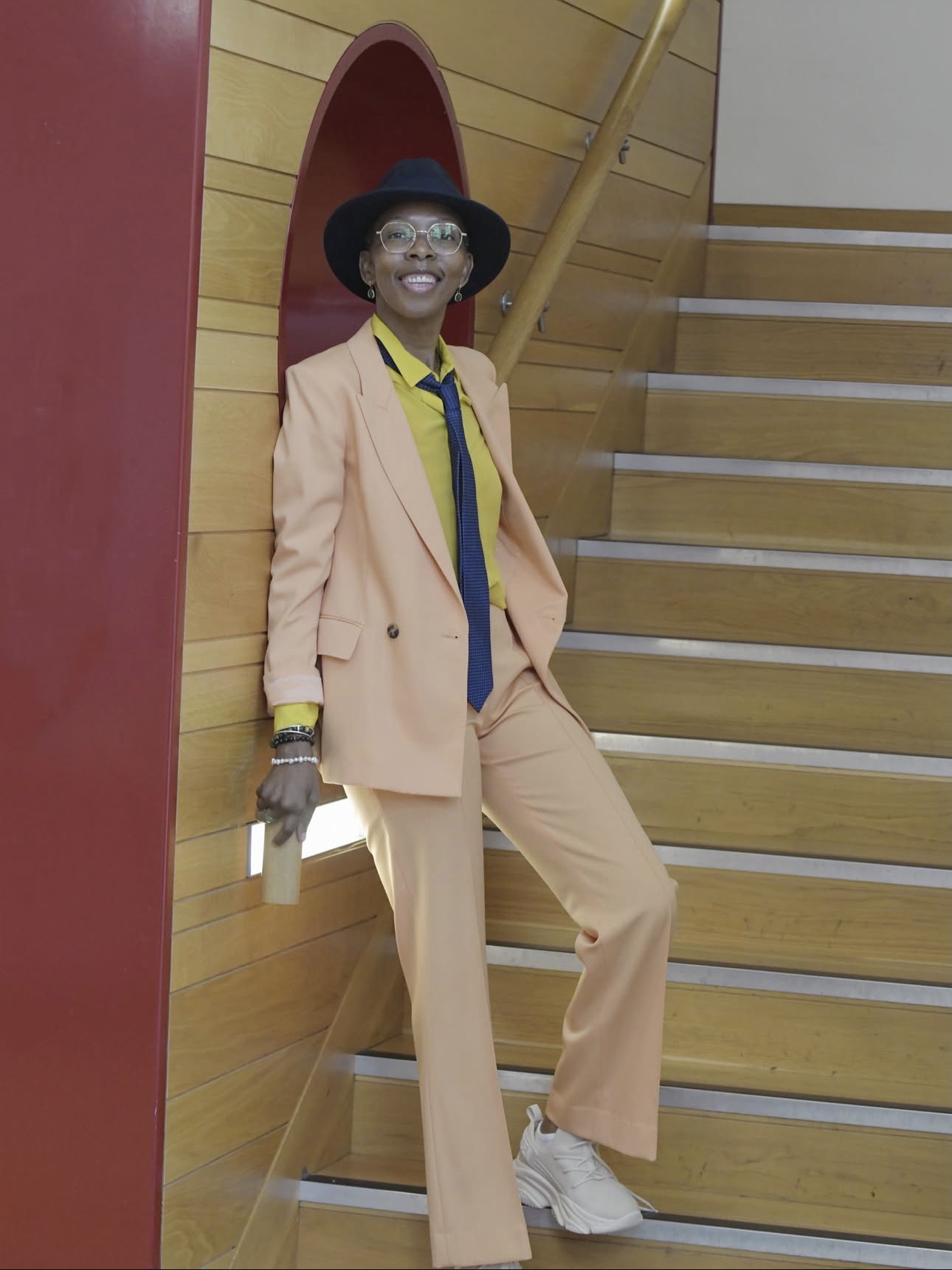 Lily Samuels smiling, in a peach suit and navy hat, leaning against a wooden staircase